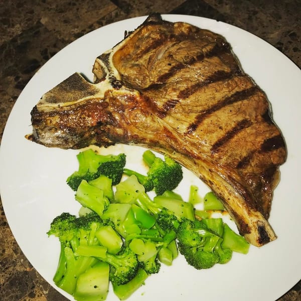 Grilled T-bone steak with prominent grill marks, served alongside steamed broccoli florets on a white plate.
