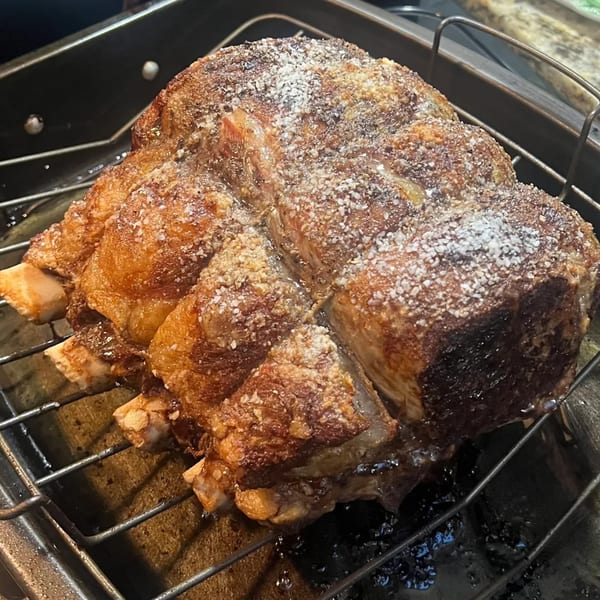 A large, seasoned prime rib roast with bones sits on a metal rack in a roasting pan.