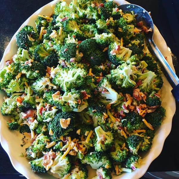 A large white platter filled with broccoli salad, topped with shredded cheese and bacon bits.