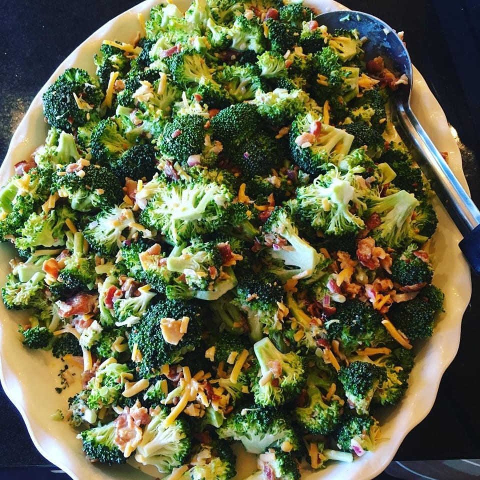 A large white platter filled with broccoli salad, topped with shredded cheese and bacon bits.